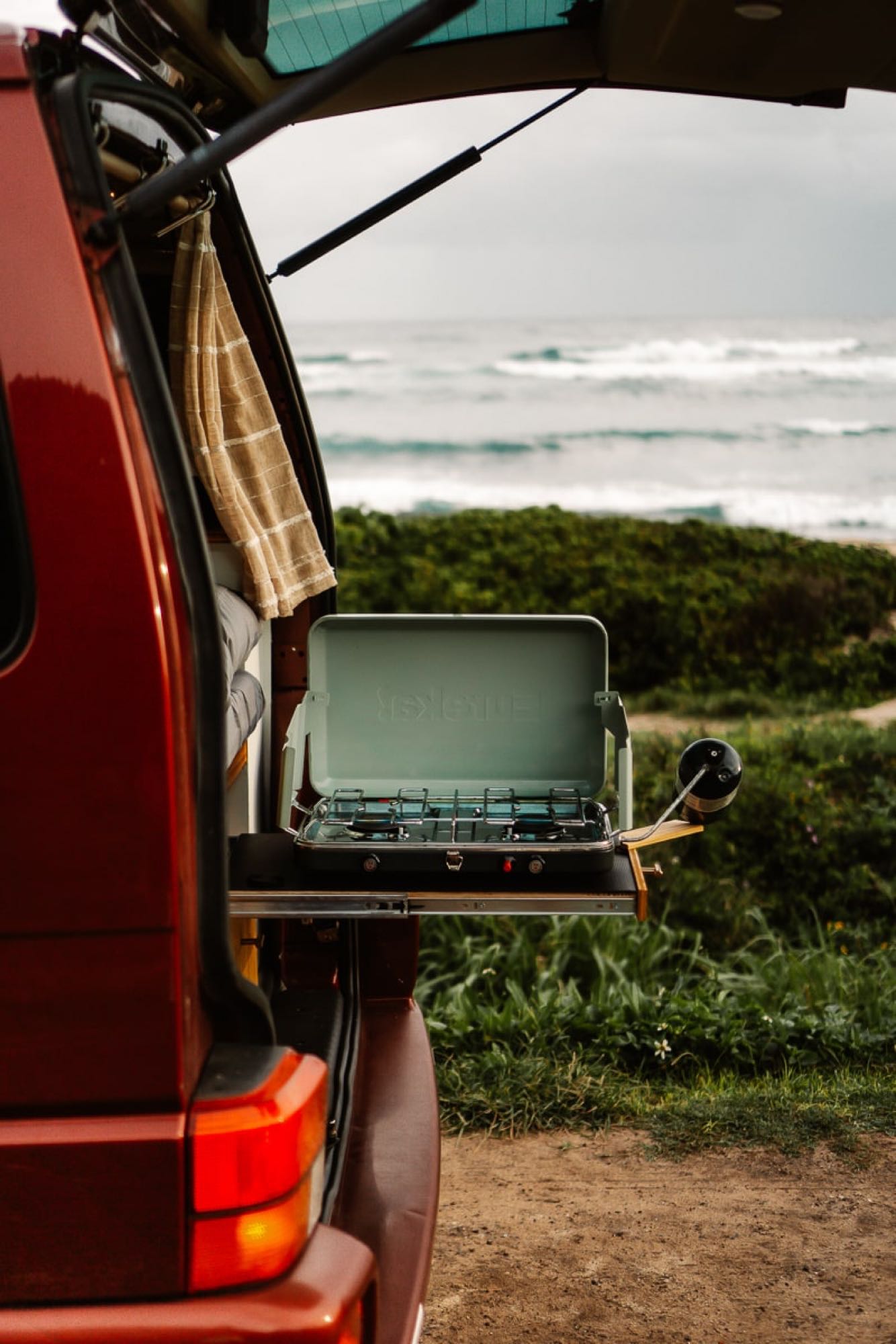 Enjoy outdoor cooking with this rv rental! A camper van's pull-out stove with ocean waves in the background.