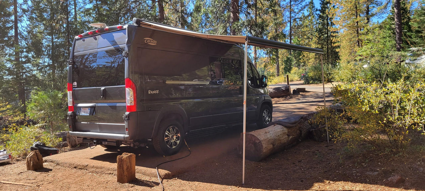 Black RV rental with extended awning parked in a sunny forest campsite, ideal for nature adventures. — photo 12