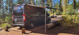Black RV rental with extended awning parked in a sunny forest campsite, ideal for nature adventures.