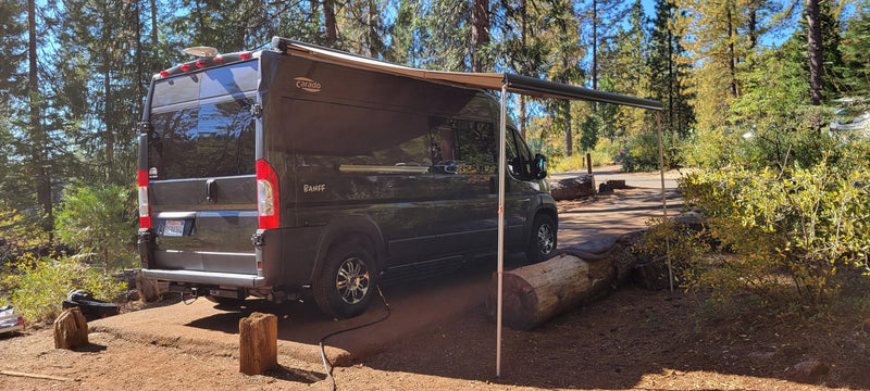 Black RV rental with extended awning parked in a sunny forest campsite, ideal for nature adventures. — photo 12