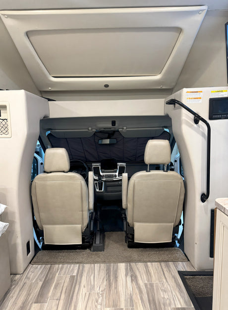 Interior view of an RV rental, showing the cockpit with two tan seats, dashboard, and an overhead bunk/storage area.