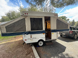 Pop-up camper RV rental set up with open tent sides and interior access, hitched to a gray SUV in an outdoor setting.