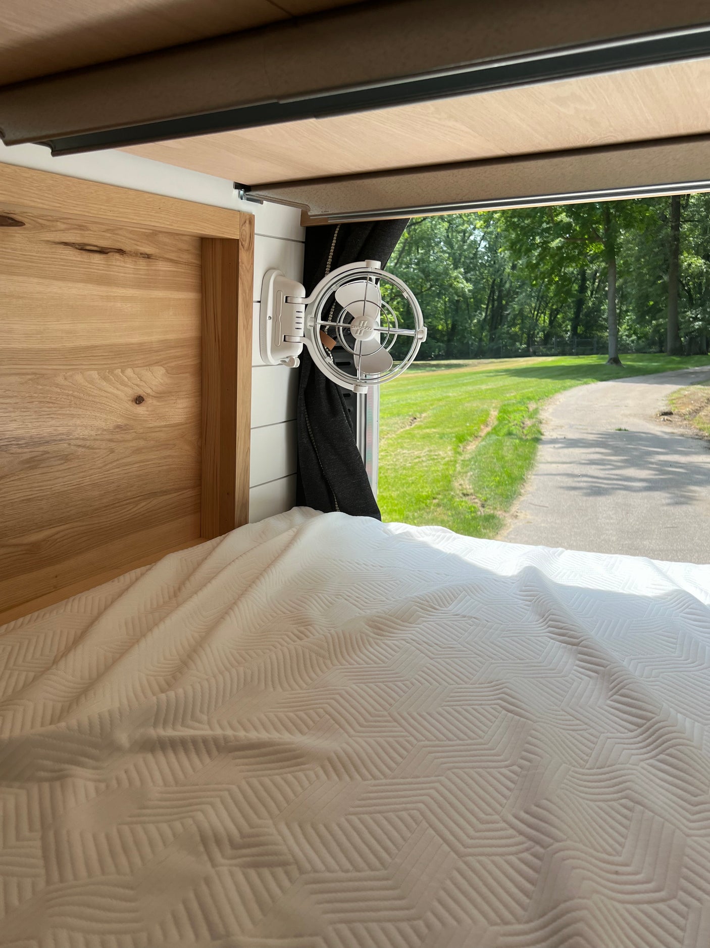 Inside a modern RV rental, featuring a comfortable bed with a patterned comforter, wall-mounted fan, and a serene window view of nature.