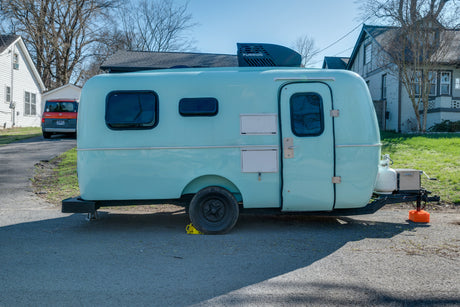 A light blue, rounded teardrop RV rental is parked on asphalt, showing its Furrion AC unit and propane tank.