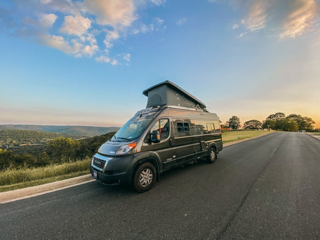 A dark gray RV rental with a pop-top roof is parked on a road, overlooking a scenic valley of green hills at sunset, under a blue sky with a crescent moon.