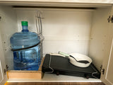 RV rental kitchen cabinet setup: large water bottle, portable griddle, pan, and spatula for camping.