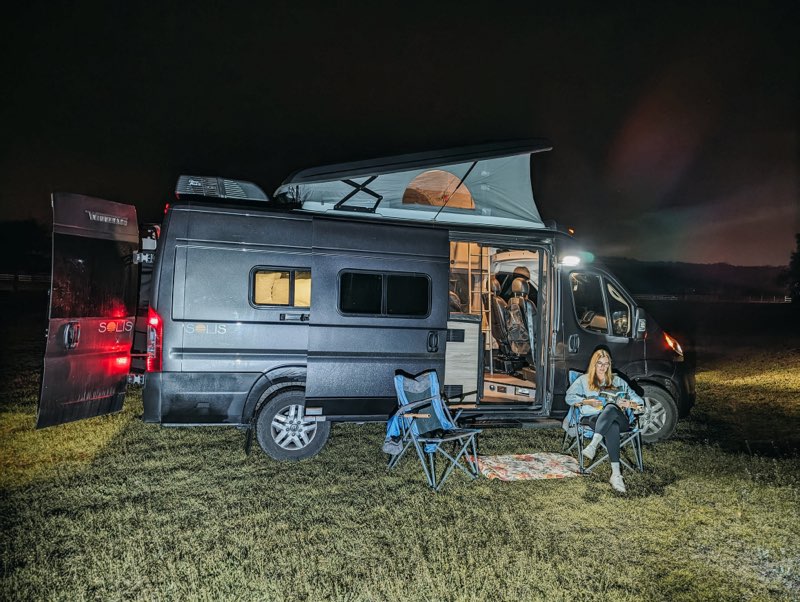 Nighttime rv rental camping: A woman enjoys reading outside her illuminated camper van in a grassy field, showcasing van life.