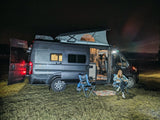 Nighttime rv rental camping: A woman enjoys reading outside her illuminated camper van in a grassy field, showcasing van life.