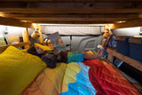 A child lies on a rainbow-colored bed, reading a book inside a well-lit RV rental, offering a cozy retreat.