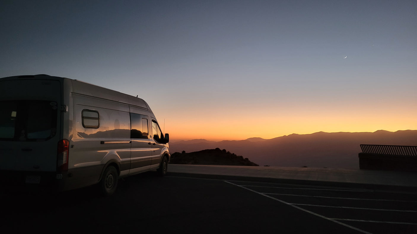 An RV rental van parked at a scenic mountain overlook during a vivid sunset, with a crescent moon above.