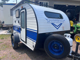 White and blue Classic Sunray teardrop camper, ideal for RV rental, parked outdoors with propane tank & spare tire.