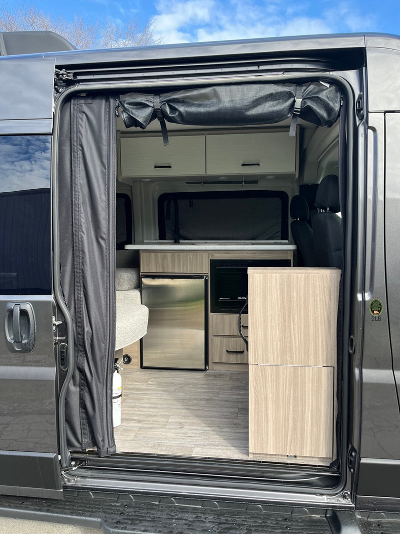 Open side door of an RV rental, showing its light-toned kitchen, fridge, microwave, and privacy curtain. — photo 13