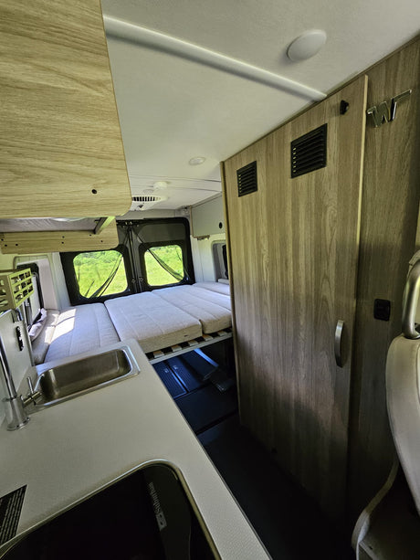Modern RV rental interior: compact camper van features a convertible bed, kitchen sink, stove, and light wood cabinetry.