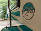 A distinctive white and teal T@G RV rental, featuring its unique metallic logo and striped side. Ideal for memorable travels.