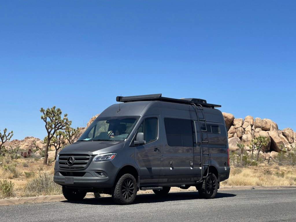 This grey Sprinter van RV rental is ready for desert adventures, parked among Joshua trees and rock formations.