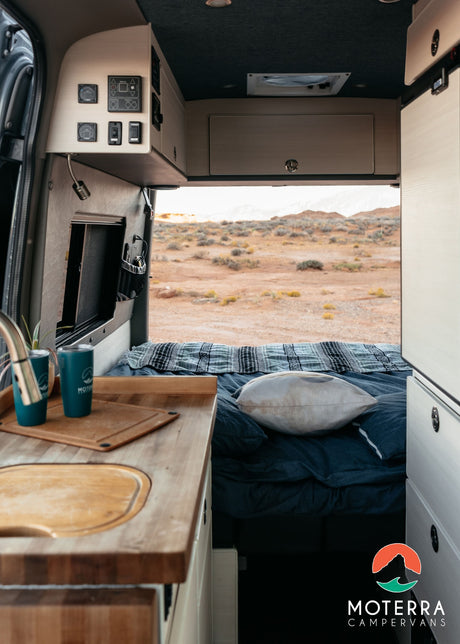 RV rental interior: cozy bed, kitchen counter, and open rear doors with a desert landscape view. Perfect for your campervan adventure.