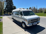 White Roadtrek RV rental (camper van) with high roof, parked on a sunny street next to a sidewalk. Ready for adventure.
