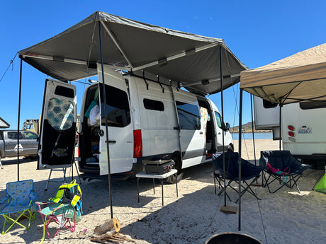 White RV rental camper van with awning, chairs, and grill set up on a sandy beach by the water, perfect for outdoor living.