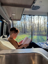 A young boy reads in a cozy **RV rental** bed, enjoying sunlit woods outside. Perfect for a relaxing travel moment.