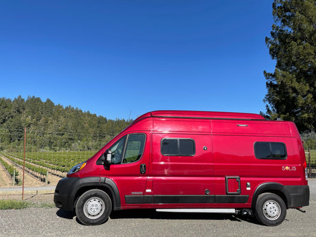Red Winnebago Solis camper van for RV rental, parked by a sunny vineyard and forest hills under a clear blue sky.