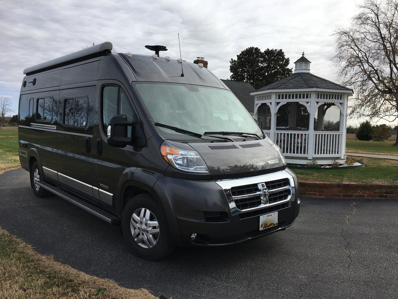  and dark grey exterior.|Sleek dark grey Winnebago Travato RV rental parked on asphalt with a house and bare trees in the background.|A grey Winnebago RV van is parked on asphalt beside a grassy slope leading to a calm bay under a cloudy sky — photo 4