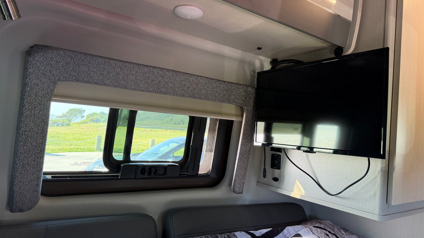 RV rental interior: window with green landscape view, wall-mounted TV, and bed/seating with patterned fabric.