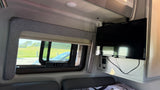 RV rental interior: window with green landscape view, wall-mounted TV, and bed/seating with patterned fabric.