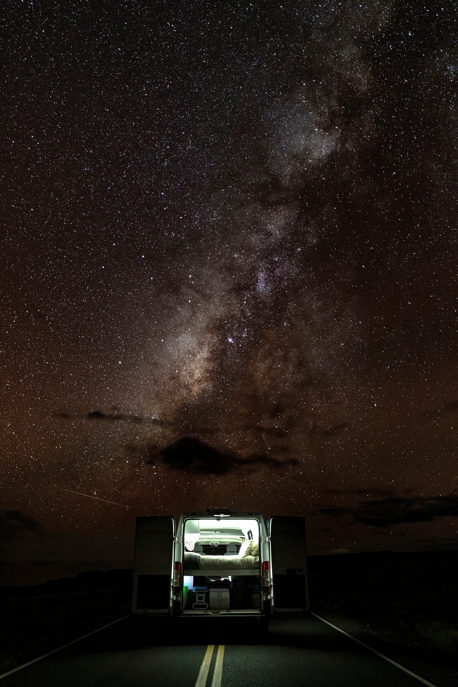 An RV rental camper van, lit from inside, parked on a dark road under a vast, star-filled Milky Way. Perfect for night sky camping.