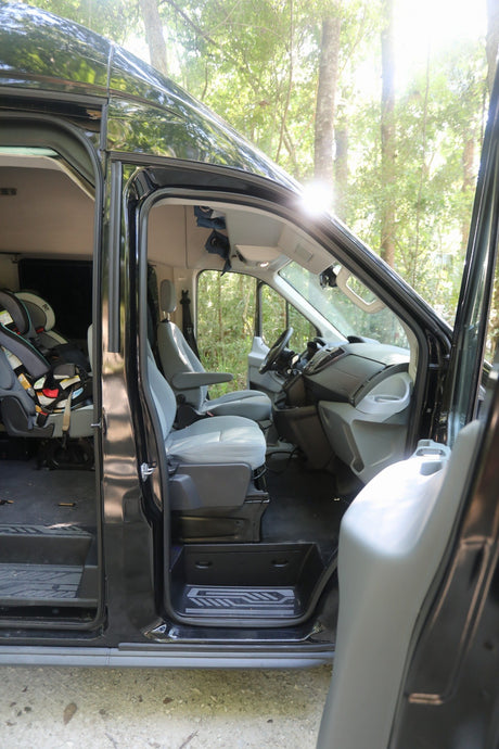 RV rental van interior: child safety seats visible, doors open in a sunny forest.
