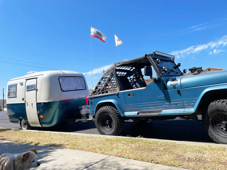 A blue Jeep tows a compact, retro RV trailer under California flags. Ready for adventure, this RV rental is perfect for camping. Dog on sidewalk.