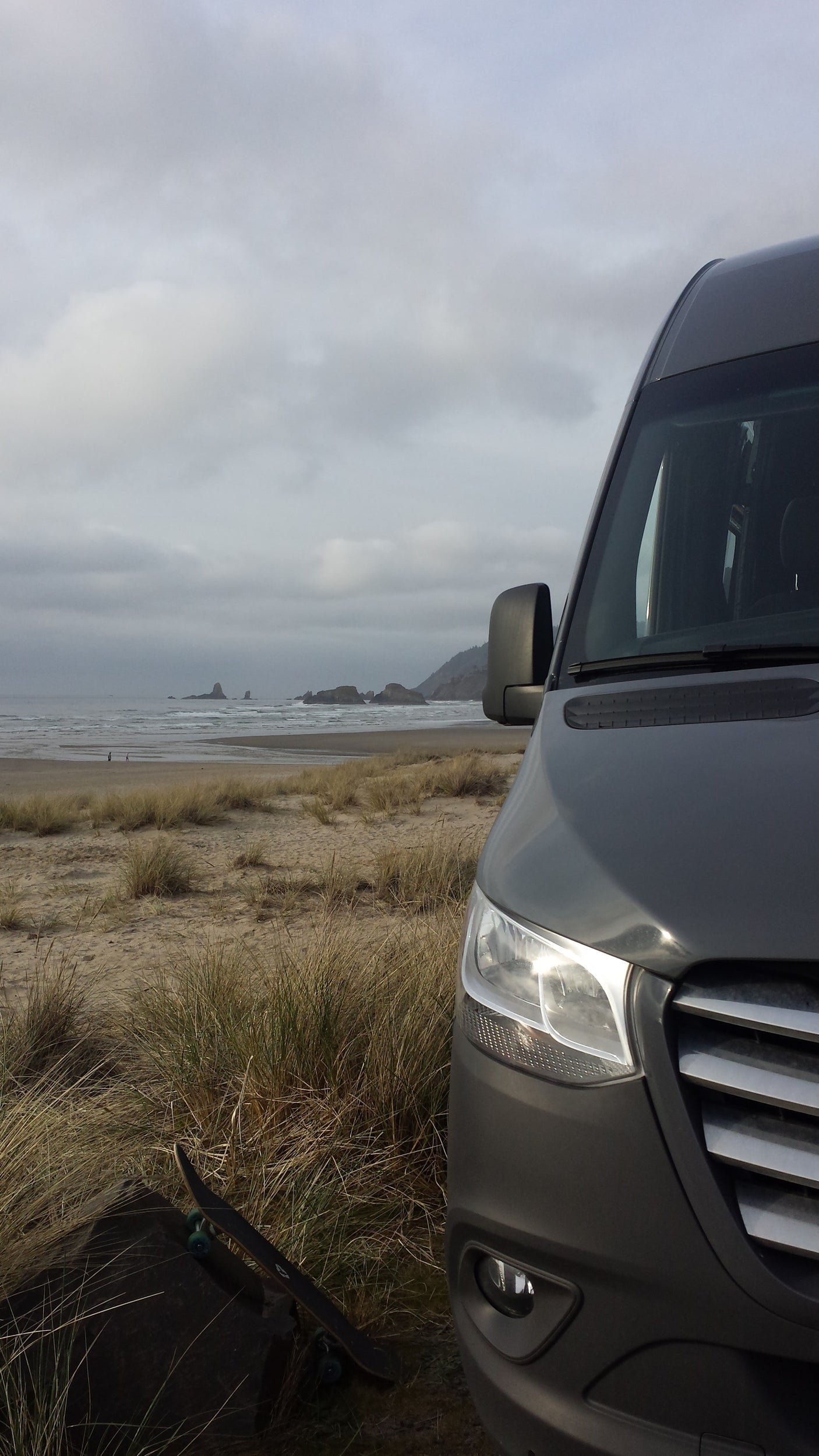 A grey RV rental van parked on a sandy beach with tall grass, facing ocean waves, distant sea stacks, and a skateboard nearby.
