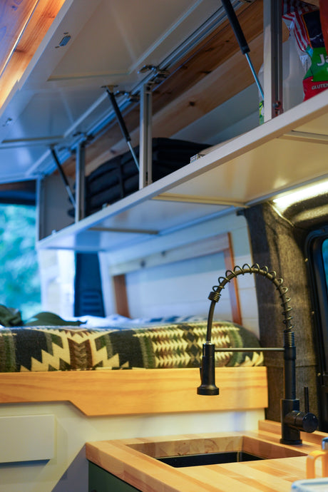 Inside a modern RV rental: kitchen with sleek faucet, wood counter, and overhead cabinets above a comfy bed.