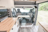 Inside a modern rv rental van: kitchenette, swivel front seats with table setup. Views of water & greenery.