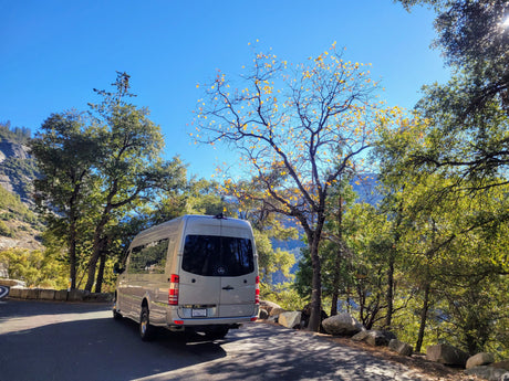 Silver Mercedes-Benz Sprinter RV rental parked facing away on a mountain road, with autumn trees and rock cliffs under a bright blue sky.
