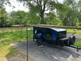 A compact blue Hiker camper, an ideal RV rental, is parked with its awning extended by a green riverbank for serene outdoor camping.