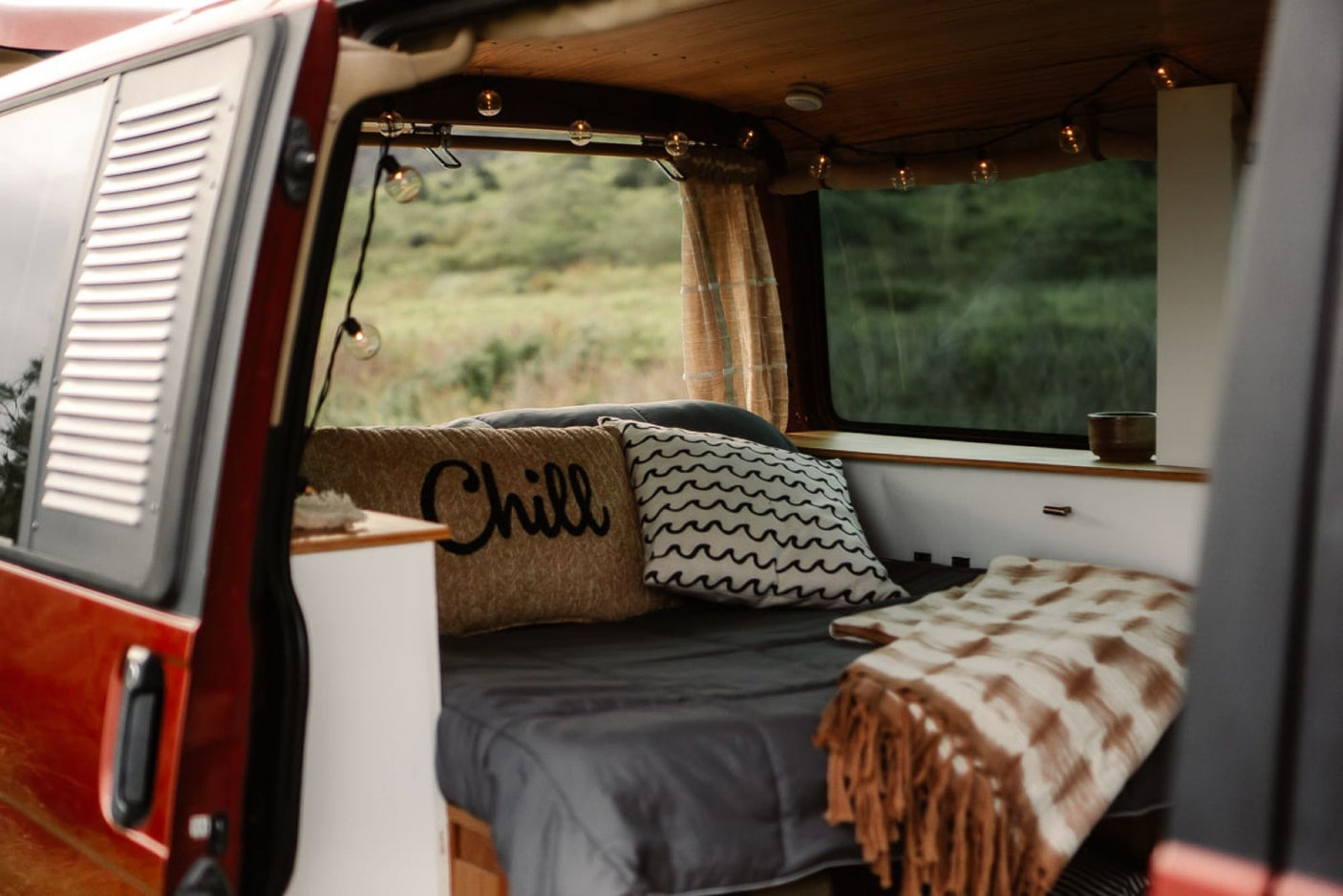 Comfortable RV rental interior with a bed, 'Chill' pillow, string lights & a window view of nature. Perfect for van life.