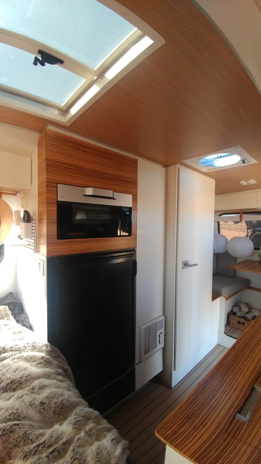 Bright RV rental interior: sleek kitchen with microwave/fridge, comfy bed with furry blanket, wood-paneled ceiling & skylight. — photo 4