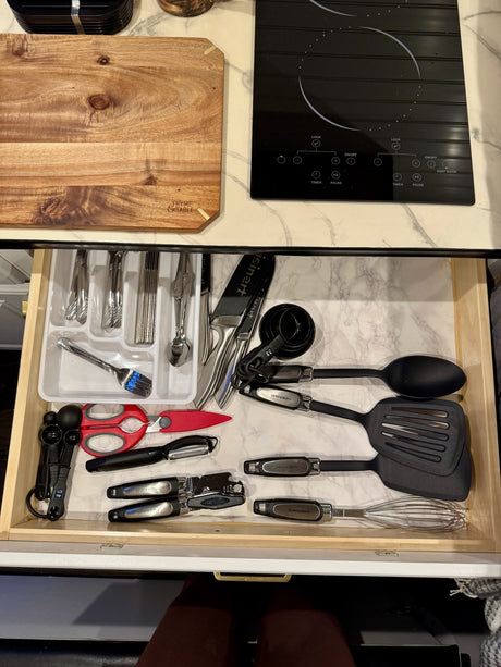 RV rental kitchen setup: Open drawer with organized utensils (spatulas, whisk, measuring spoons); countertop has induction cooktop & cutting board.