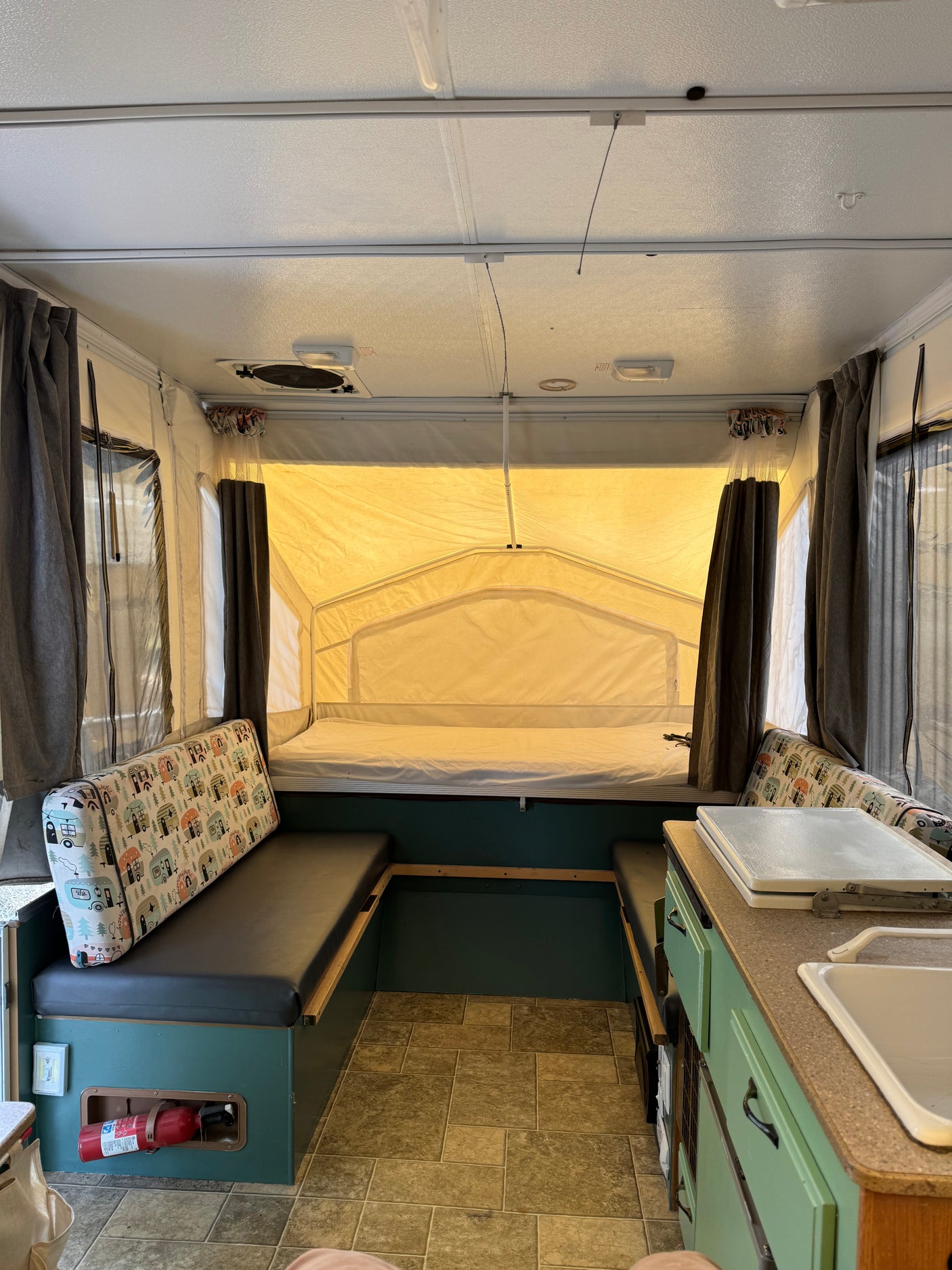 Inside a bright RV rental pop-up camper: seating with patterned cushions, a bed, and compact kitchen area with sink & stove.