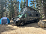 RV rental camper van with pop-top roof and blue tent at a sunny forest campsite. A large rock is nearby on the dirt ground.