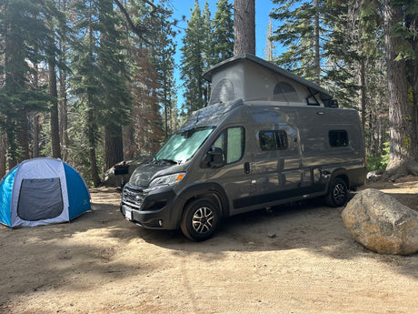 RV rental camper van with pop-top roof and blue tent at a sunny forest campsite. A large rock is nearby on the dirt ground.