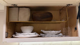 Organized RV rental kitchen cabinet features white dishware, fabric bins, wood bowl, & hanging wine glasses on a wire shelf.