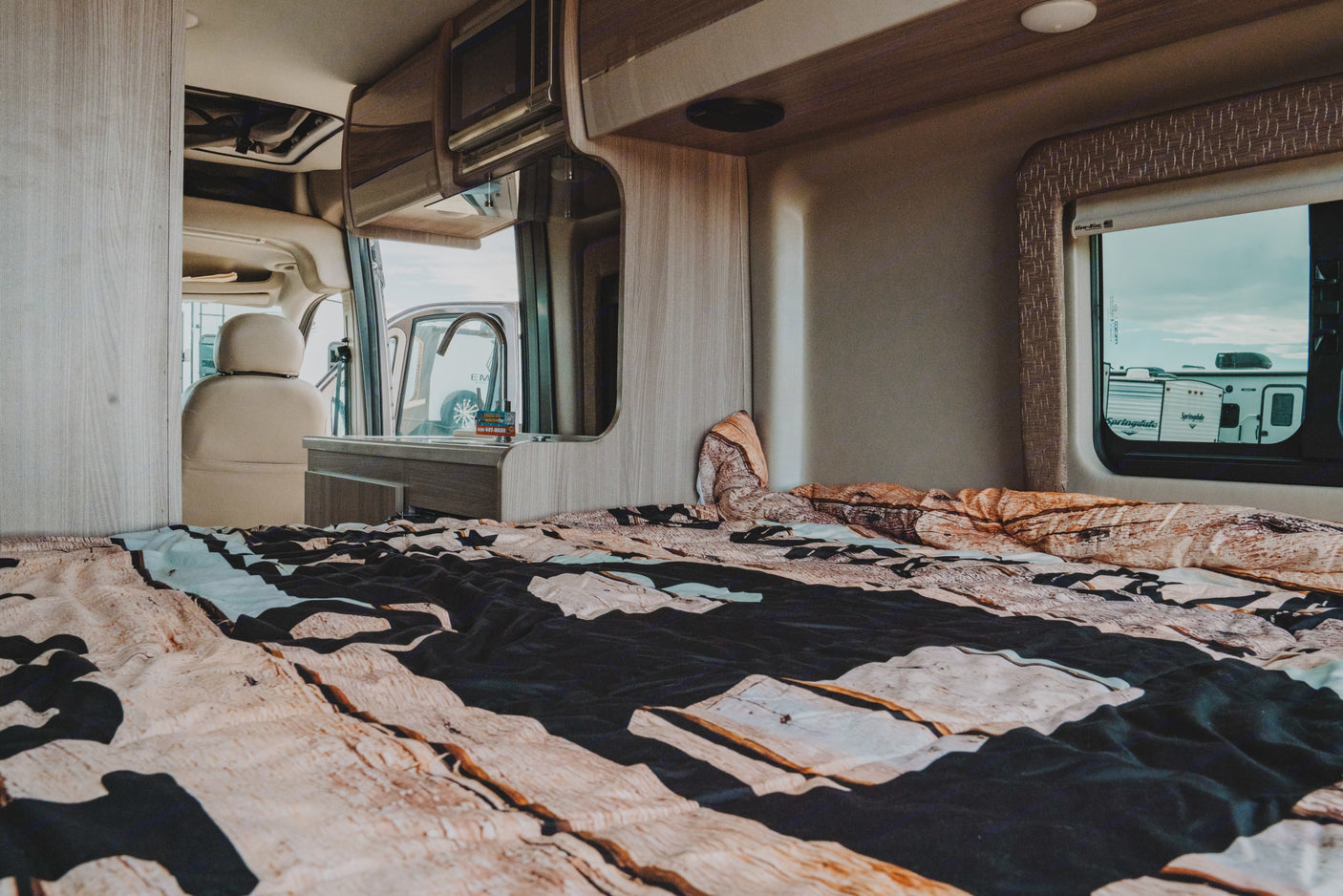 RV rental interior: bedroom with unique wood-patterned bed, kitchenette with microwave/sink, and outside view.