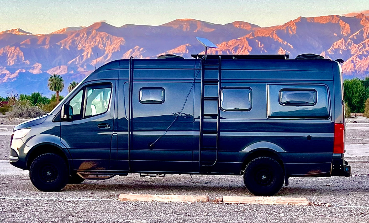 A custom dark camper van, ready for an rv rental road trip, parked against a breathtaking backdrop of sunlit desert mountains.