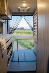 Rv rental camper van interior. A cozy table & bed setup faces open back doors with a mesh screen, revealing a beautiful field view.