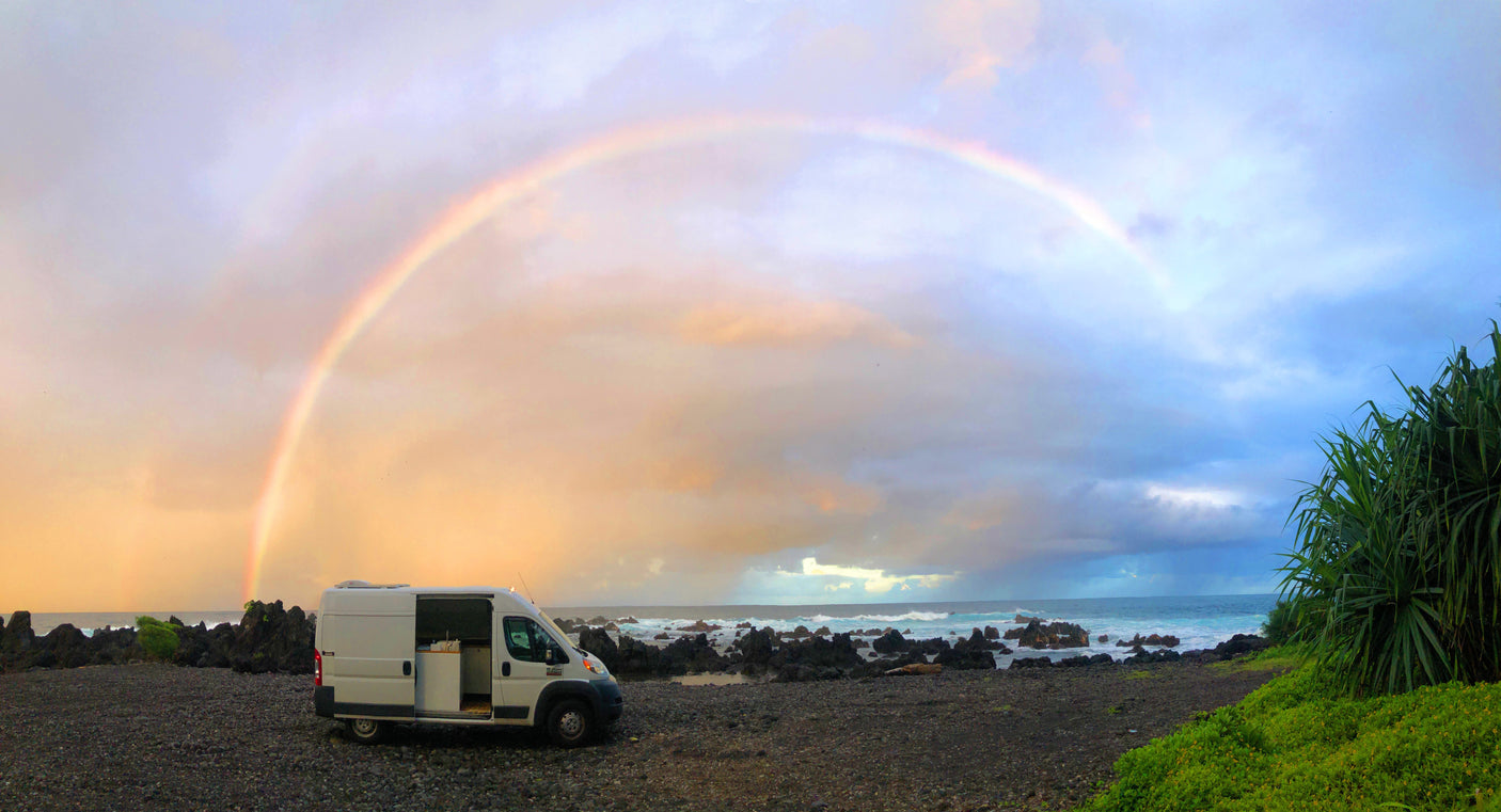 RV rental on a black beach under a vibrant rainbow. Ocean waves crash near volcanic rocks & green plants.