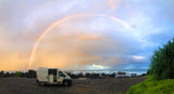 RV rental on a black beach under a vibrant rainbow. Ocean waves crash near volcanic rocks & green plants.