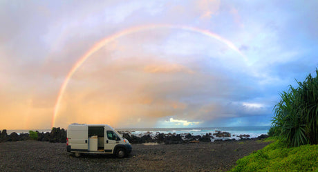 RV rental on a black beach under a vibrant rainbow. Ocean waves crash near volcanic rocks & green plants.