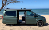 Green camper van, equipped with a Thule awning, parked on a sandy beach overlooking the ocean. Ideal for RV rental.
