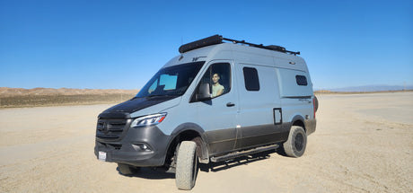 A gray Winnebago Revel RV rental, with a person in the driver's seat, sits on a wide, flat desert plain under blue skies.